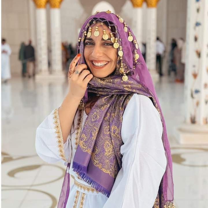 Viaggiatrice con foulard viola e monete dorate sorride nella sala di marmo bianco di una grande moschea